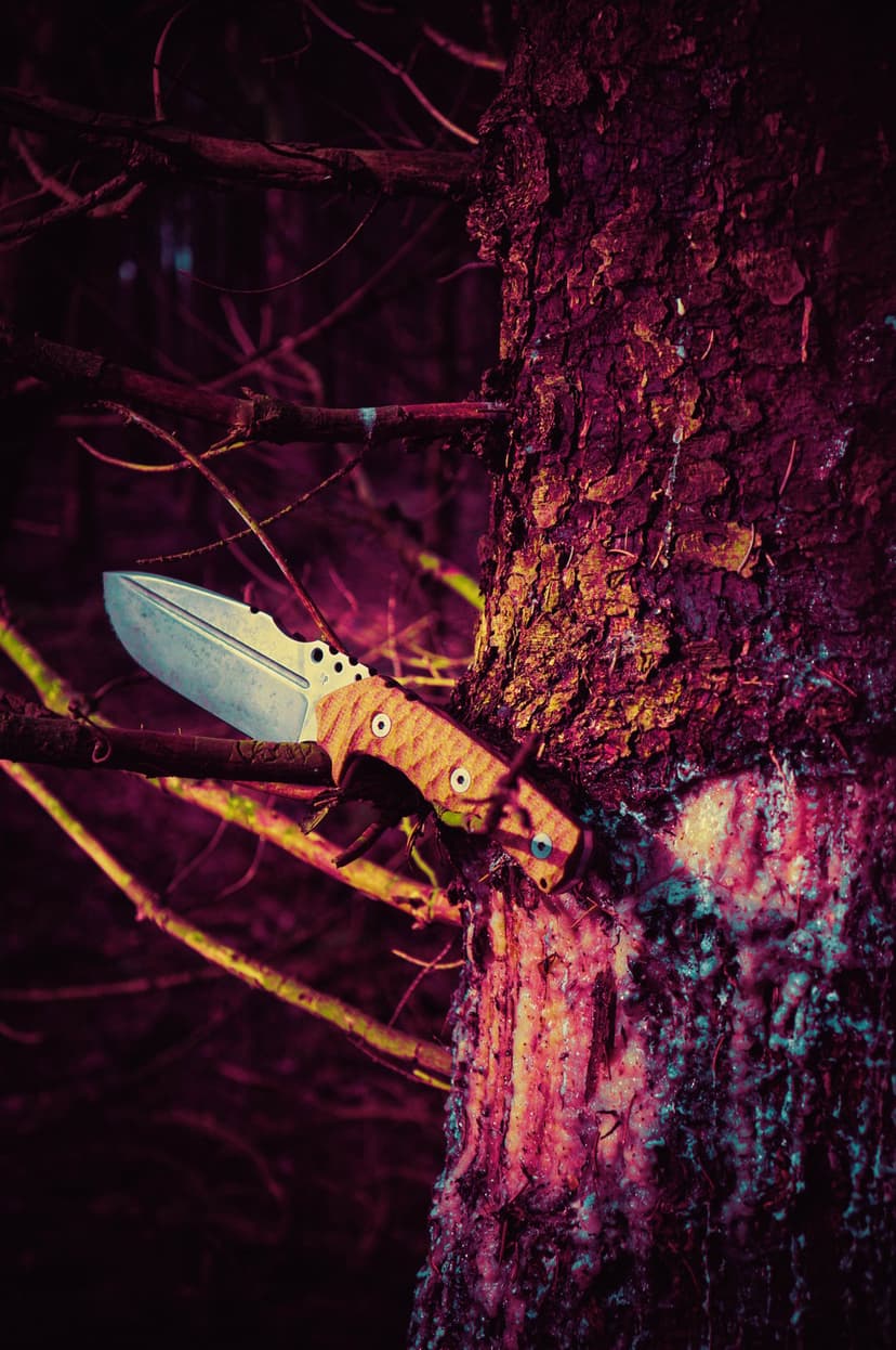 Fixed-blade knife with a wooden handle stuck into textured tree bark within a dim, moody purple forest setting.