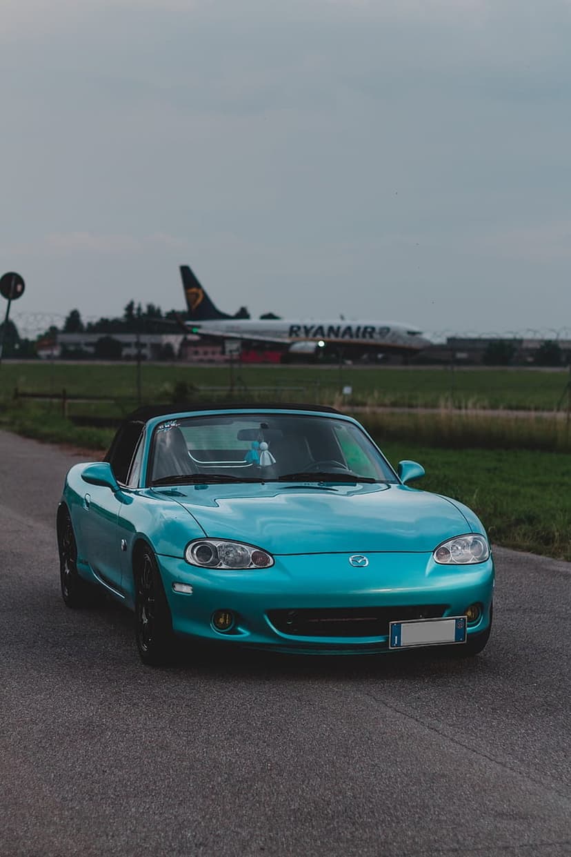 Turquoise Mazda Miata convertible parked on an asphalt road with a Ryanair jet plane on an airfield backdrop.