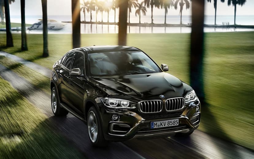 Polished black BMW X6 SUV driving along a palm-lined path toward a sunlit bay with a white yacht and palm trees.