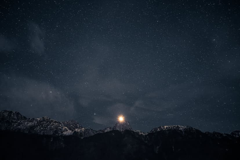 Starry night sky above silhouetted snowy mountains with a single bright beacon shining from a high rocky peak.