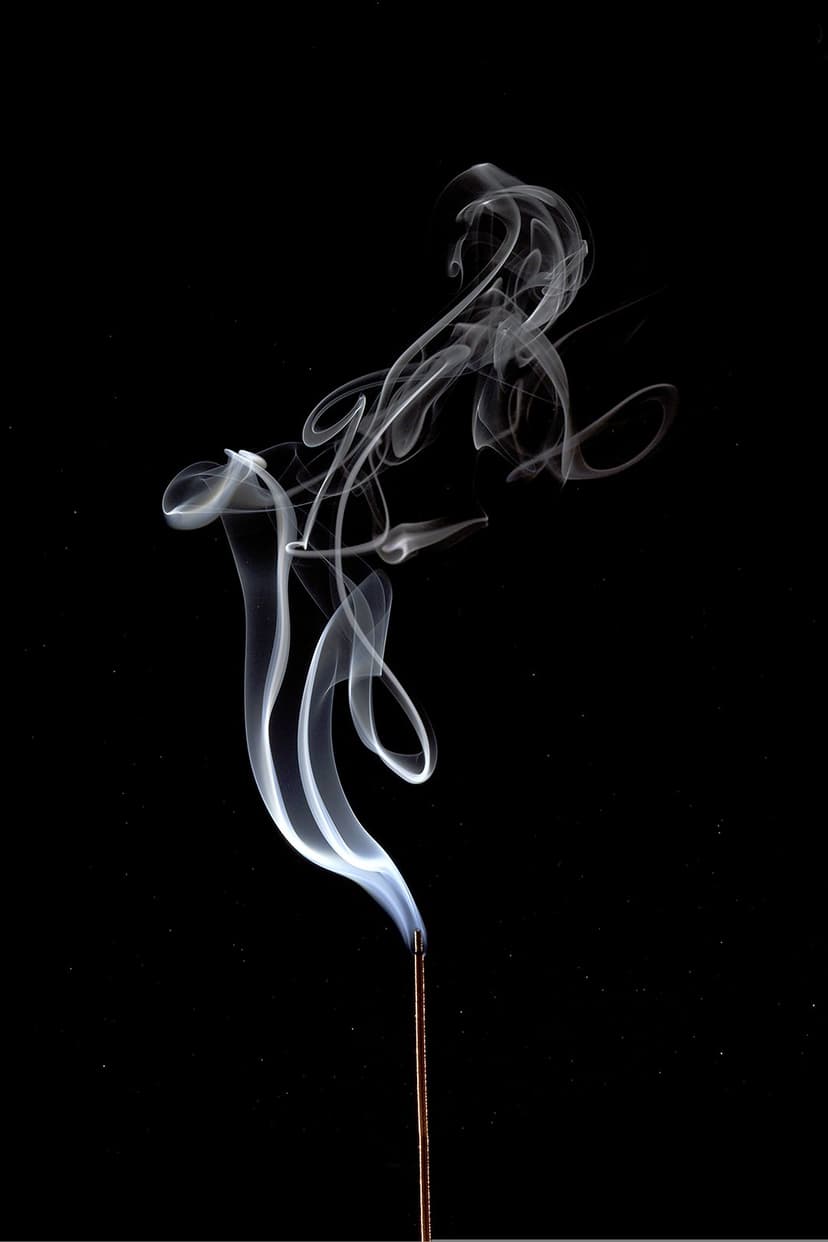 Intricate white incense smoke curls rise in delicate translucent patterns against a deep black backdrop.