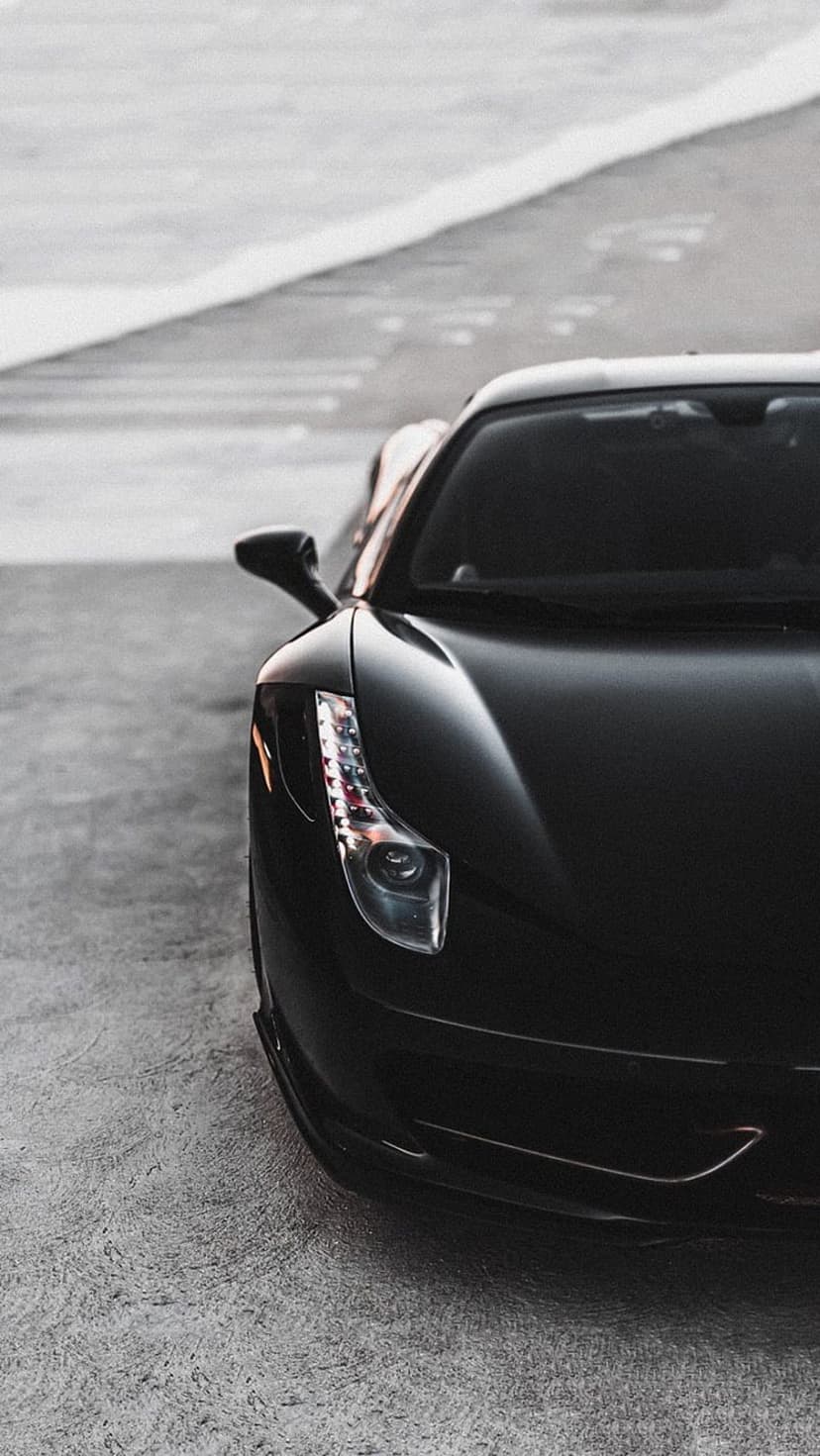 Black sports car LED headlight and front fender reflect light against a gritty urban pavement surface.
