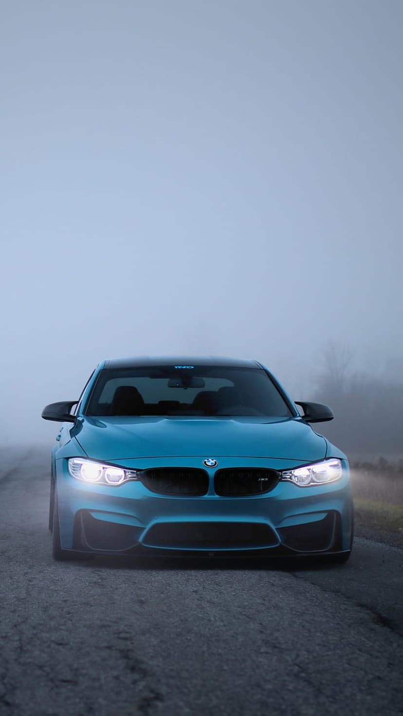 Blue BMW M4 sports car headlights shining through thick morning fog on a dark asphalt road in a vertical view.