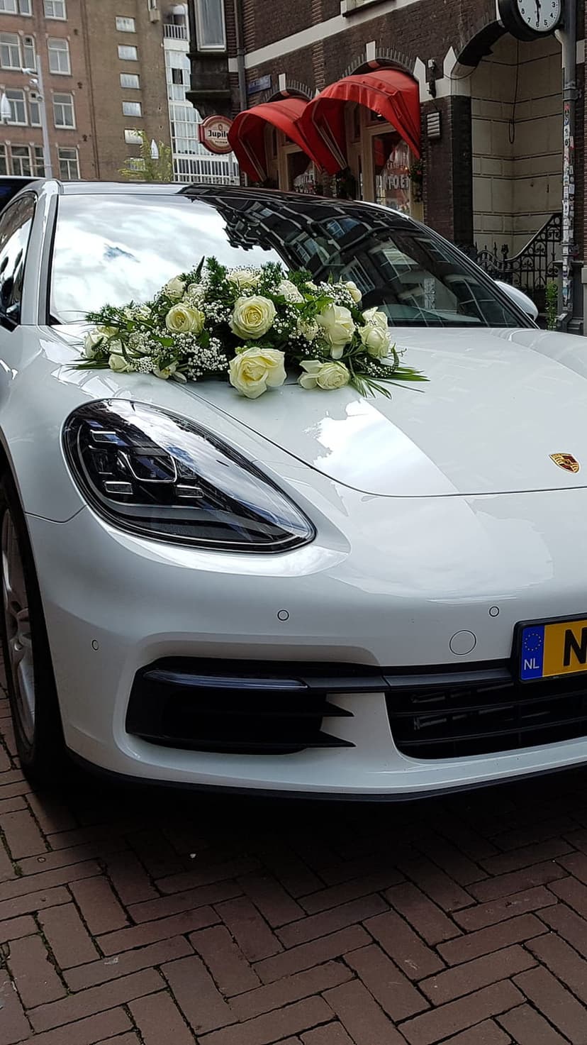 White Porsche Panamera decorated with white roses and greenery parked on a brick street during a wedding event.