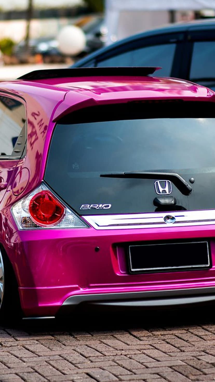 Pink Honda Brio hatchback with a custom spoiler and aftermarket rims parked on a textured brick ground.