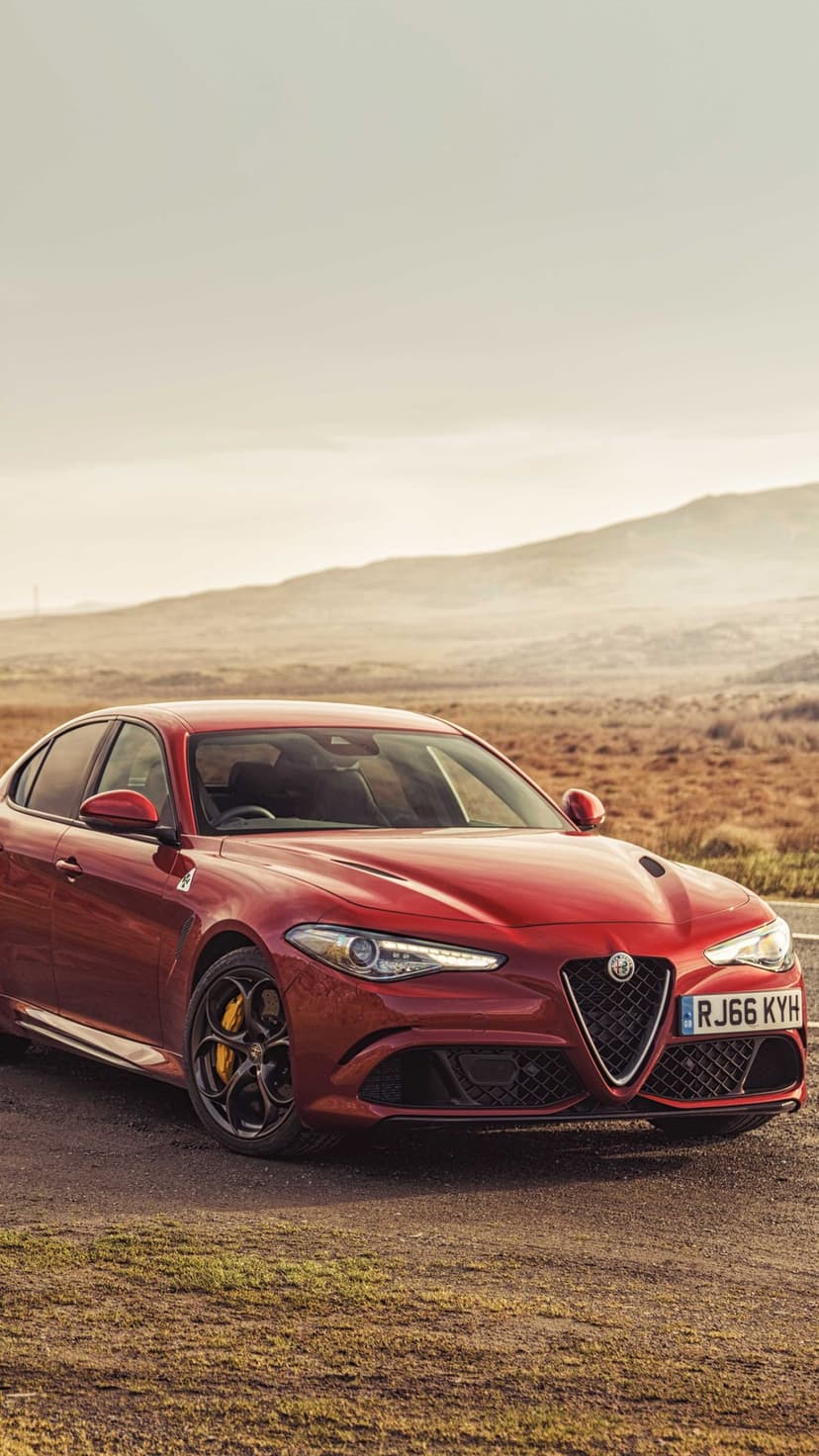 Red Alfa Romeo Giulia luxury sedan parked on a rural road against a hazy mountain landscape at sunset.