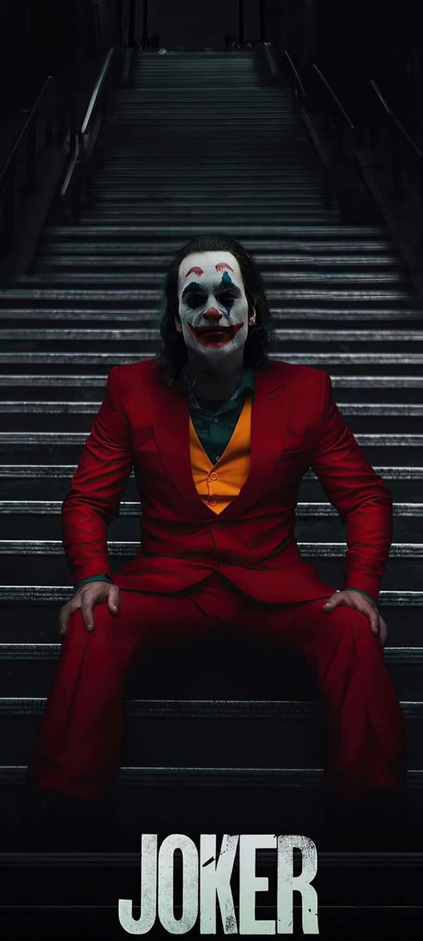 Arthur Fleck wearing a red suit and clown makeup sits on stone stairs below the bold Joker movie title.