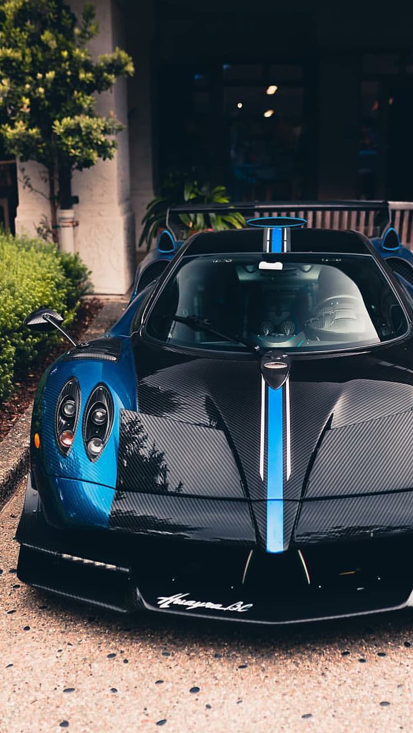 Pagani sports car with blue carbon fiber racing stripes parked near greenery, captured in a low-angle close-up.