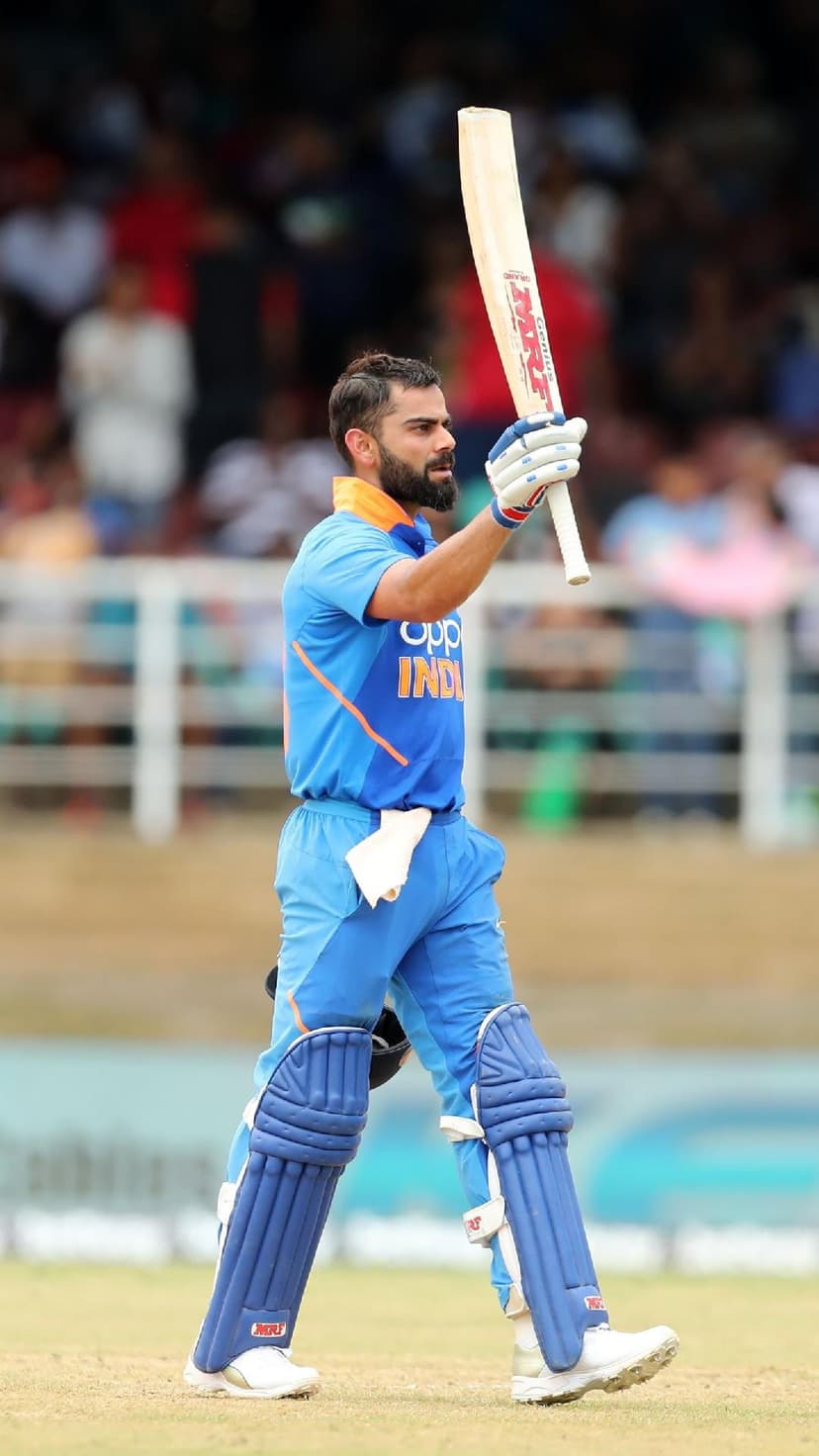 Virat Kohli raises his cricket bat in triumph while wearing a blue jersey in front of a blurred stadium crowd.