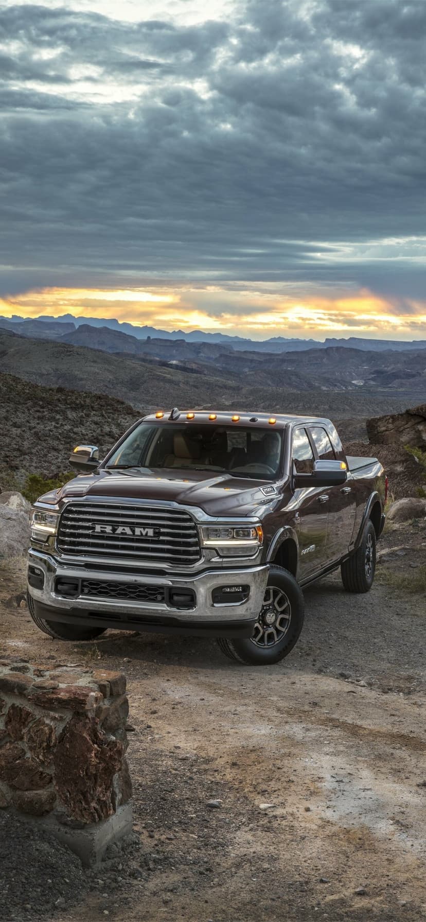 Brown Ram 2500 truck with chrome grill parked on a dirt path against a mountain sunset with cloudy sky.
