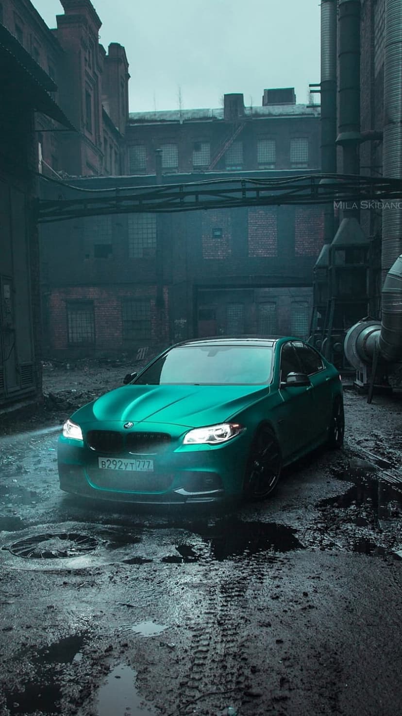 Teal BMW M5 sedan on wet ground amidst industrial brick walls and puddles, capturing a dark urban automotive scene.
