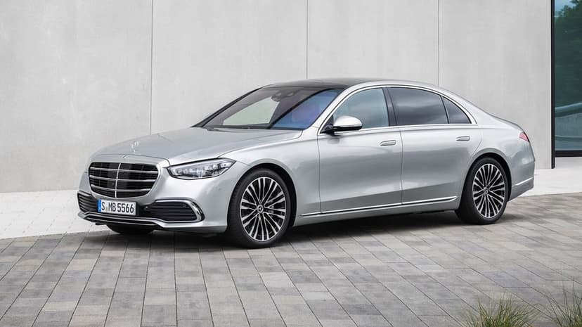 Silver Mercedes-Benz S-Class sedan parked on gray pavement against a light concrete wall background.