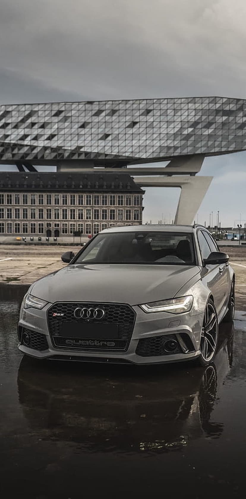 Audi RS6 Avant in gray parked on wet pavement before a faceted modern building under an overcast sky.