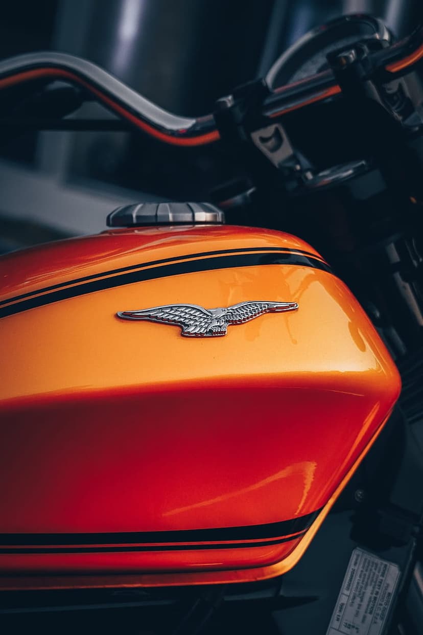 Orange motorcycle gas tank featuring black racing stripes, a silver eagle emblem, and polished chrome handlebars.