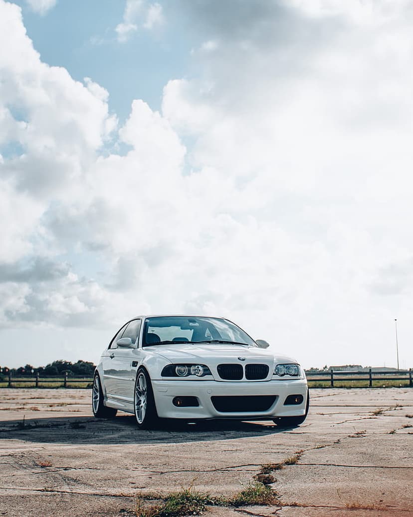 White BMW M3 E46 coupe parked on cracked concrete under a sky filled with large white clouds from a low angle.