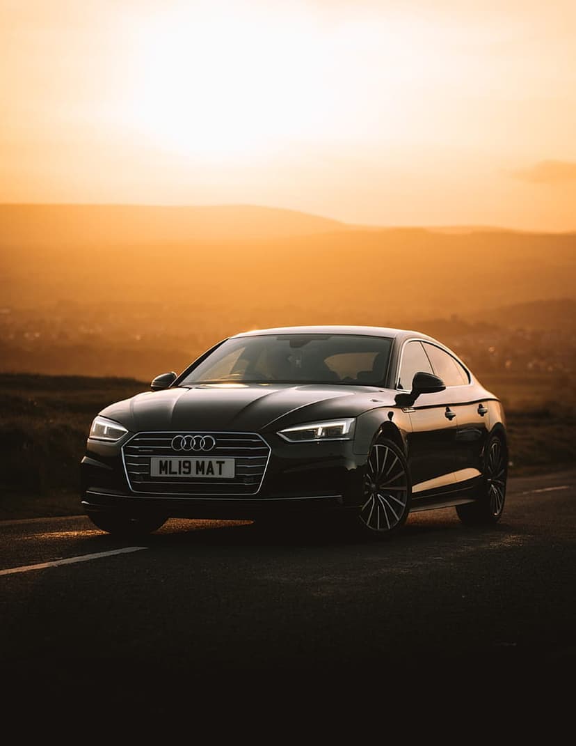 Black Audi A5 coupe with glowing headlights parked on a rural road against a vibrant orange and amber sunset.