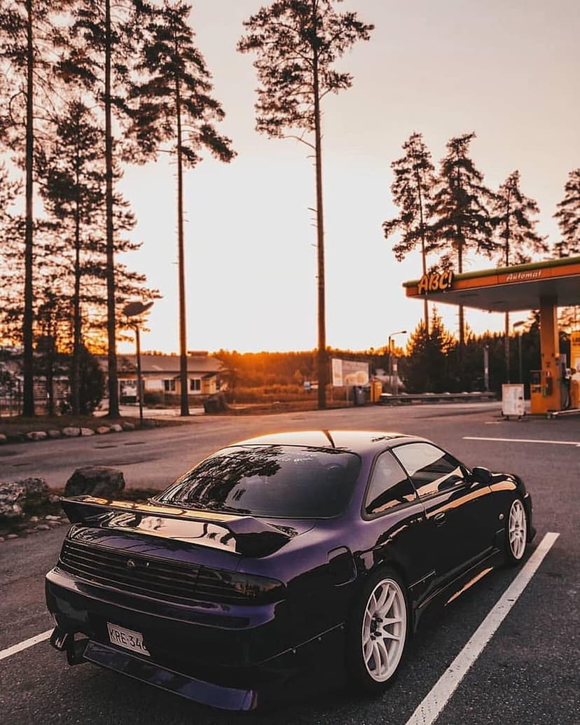 Purple Nissan S14 parked at a gas station with tall pine trees during a golden hour sunset with warm light.