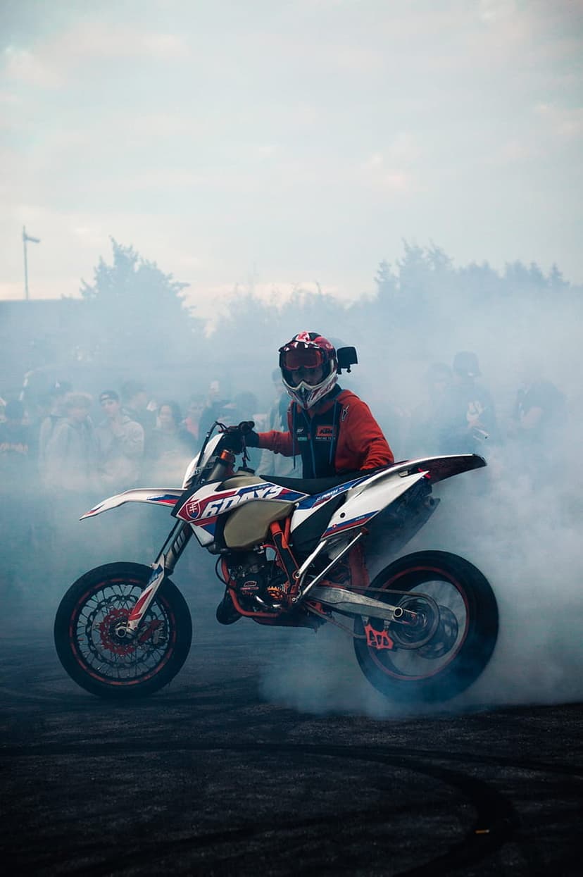 Motorcyclist in a red racing suit performs a wheelie on a white dirt bike amidst thick clouds of white smoke.
