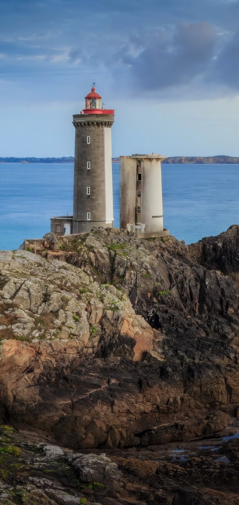 White lighthouse with a red top perches on dark rocky cliffs above a deep blue sea under a cloudy sky.