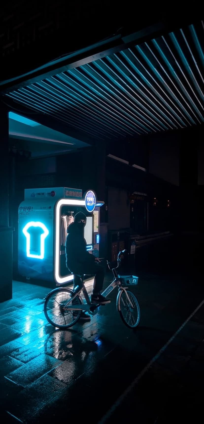 Hooded cyclist stands by a neon blue vending machine reflecting on wet pavement during a rainy night in the city.