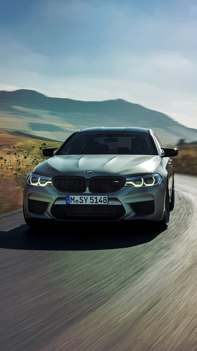 Silver BMW M5 sedan drives on a winding asphalt road with illuminated headlights against rolling hills and blue sky.
