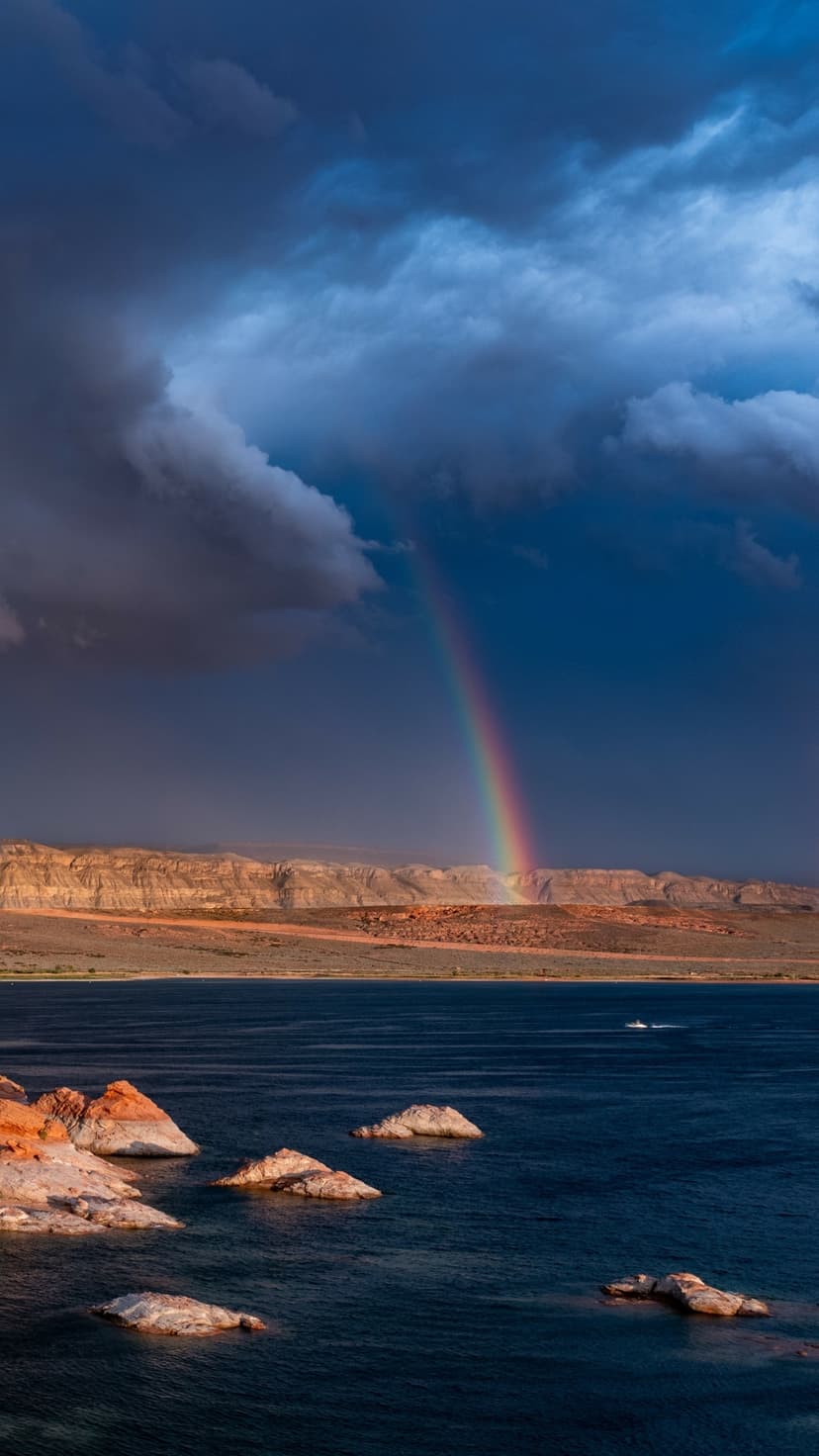 Multi-colored rainbow arches over deep blue water and red sandstone cliffs under a dark, stormy sky at Lake Powell.