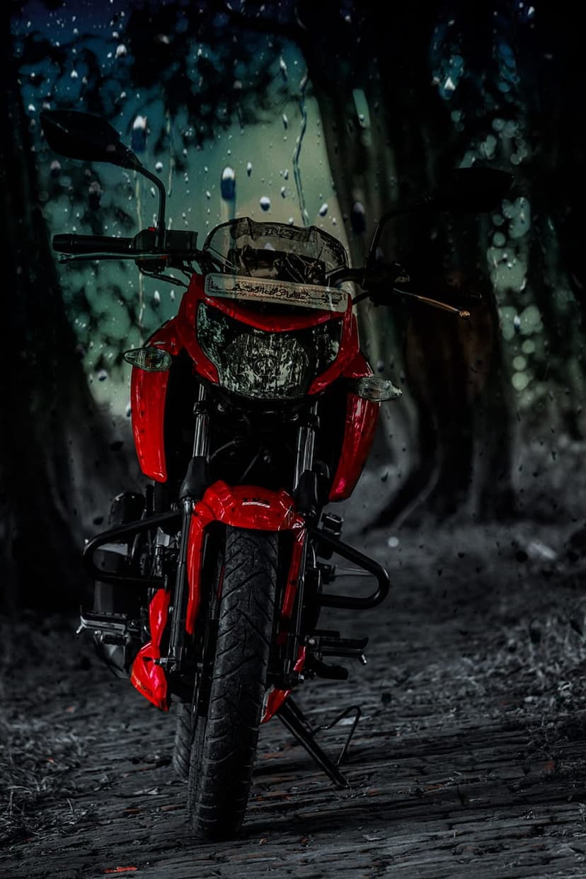 Red motorcycle parked on a weathered path inside a dark rainy forest with blurred raindrops on the camera lens.