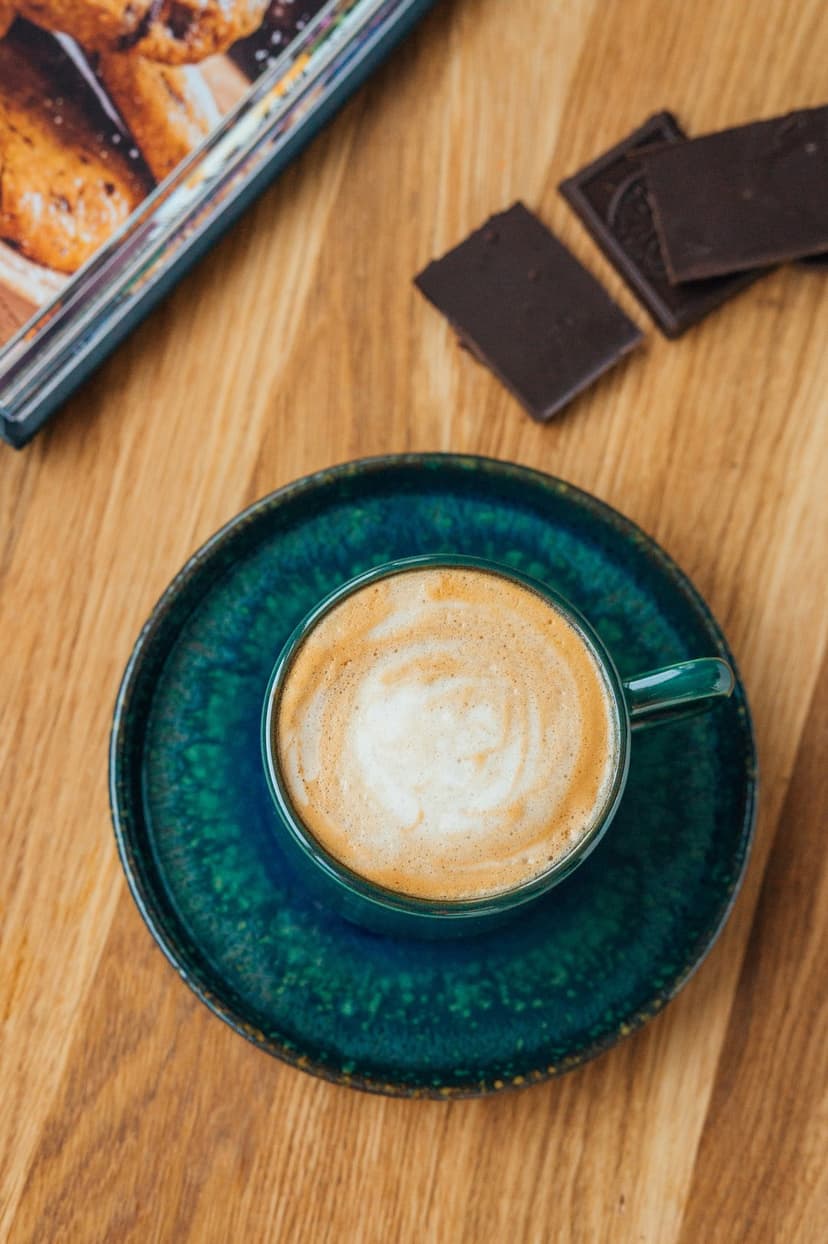 Latte art in a teal cup sits next to dark chocolate chunks on a wooden table with a blurred book nearby.