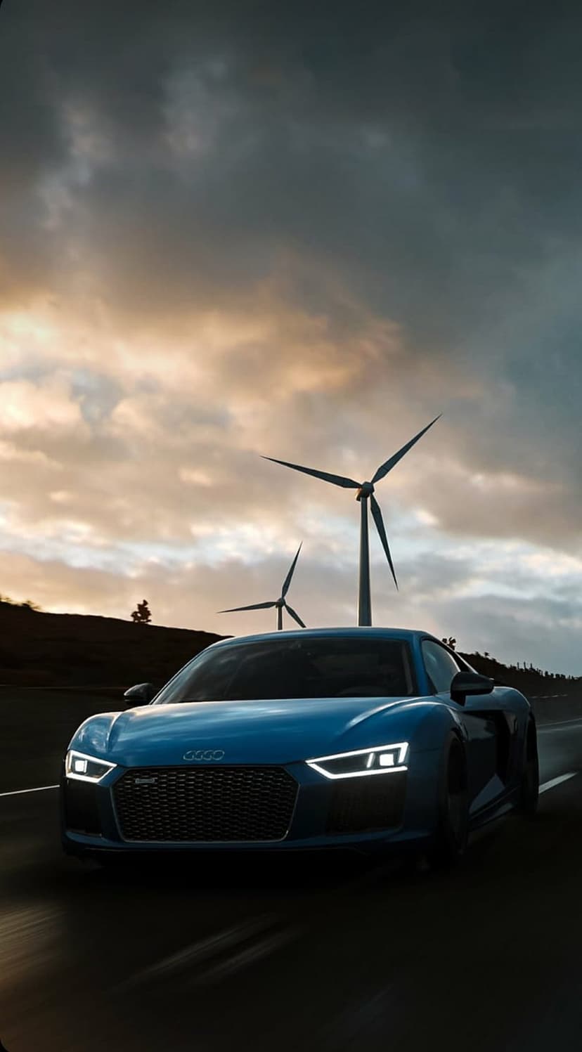 Blue Audi R8 supercar driving on an asphalt road past tall wind turbines under a dramatic orange sunset sky.