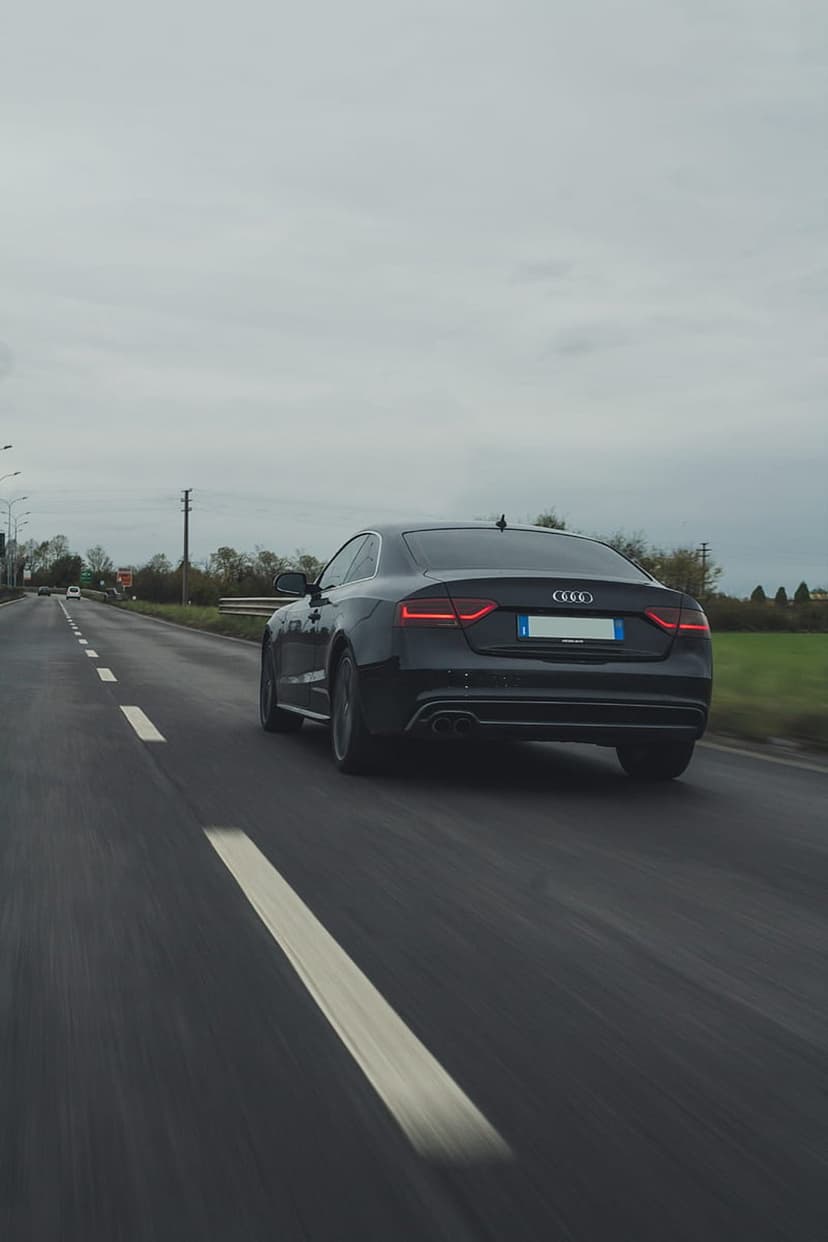 Black Audi coupe driving on an asphalt highway with motion blur under a cloudy sky, seen from the rear.