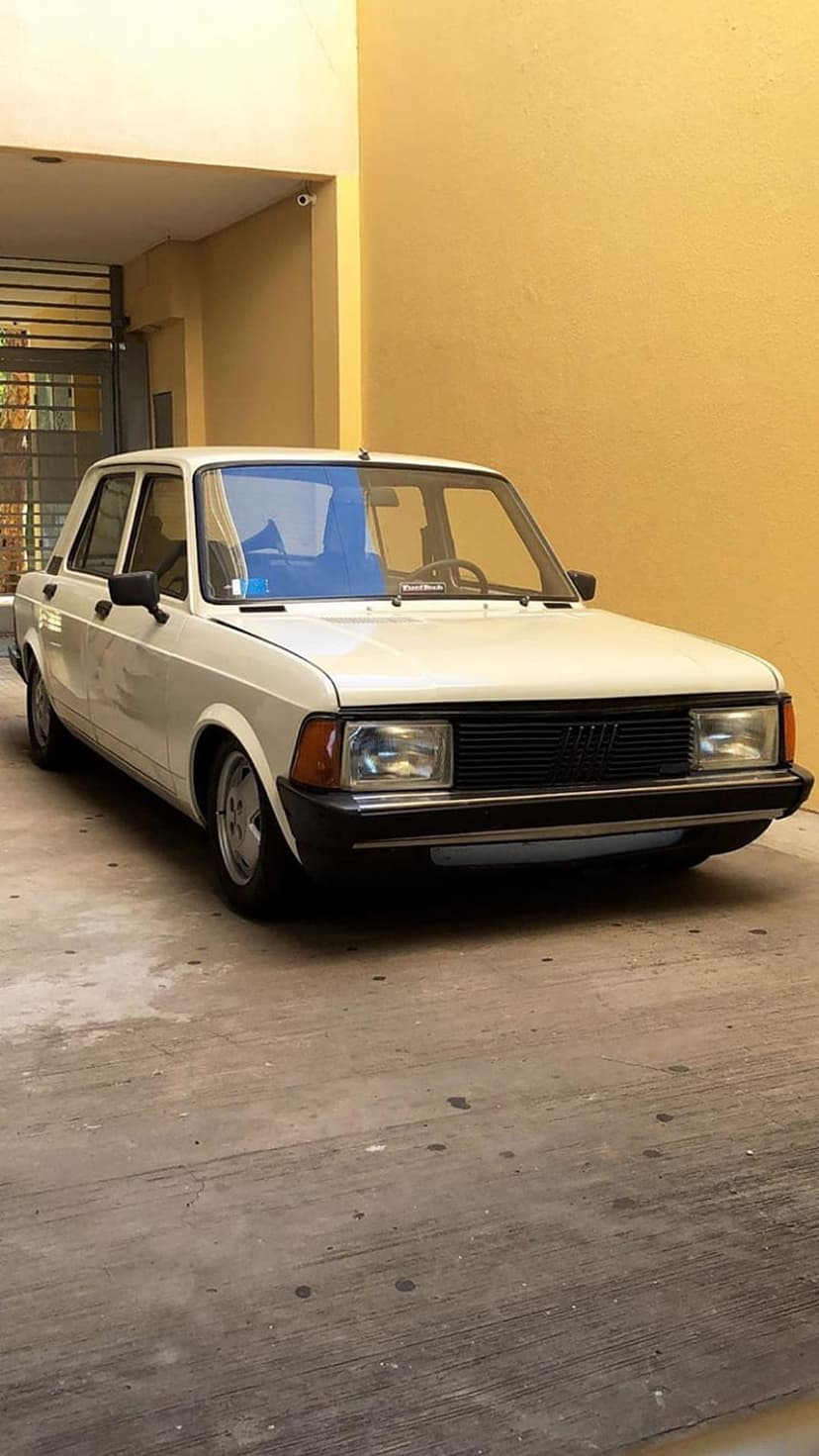 White Fiat 131 Supermirafiori sedan parked on a concrete lot beside a building with yellow exterior walls.