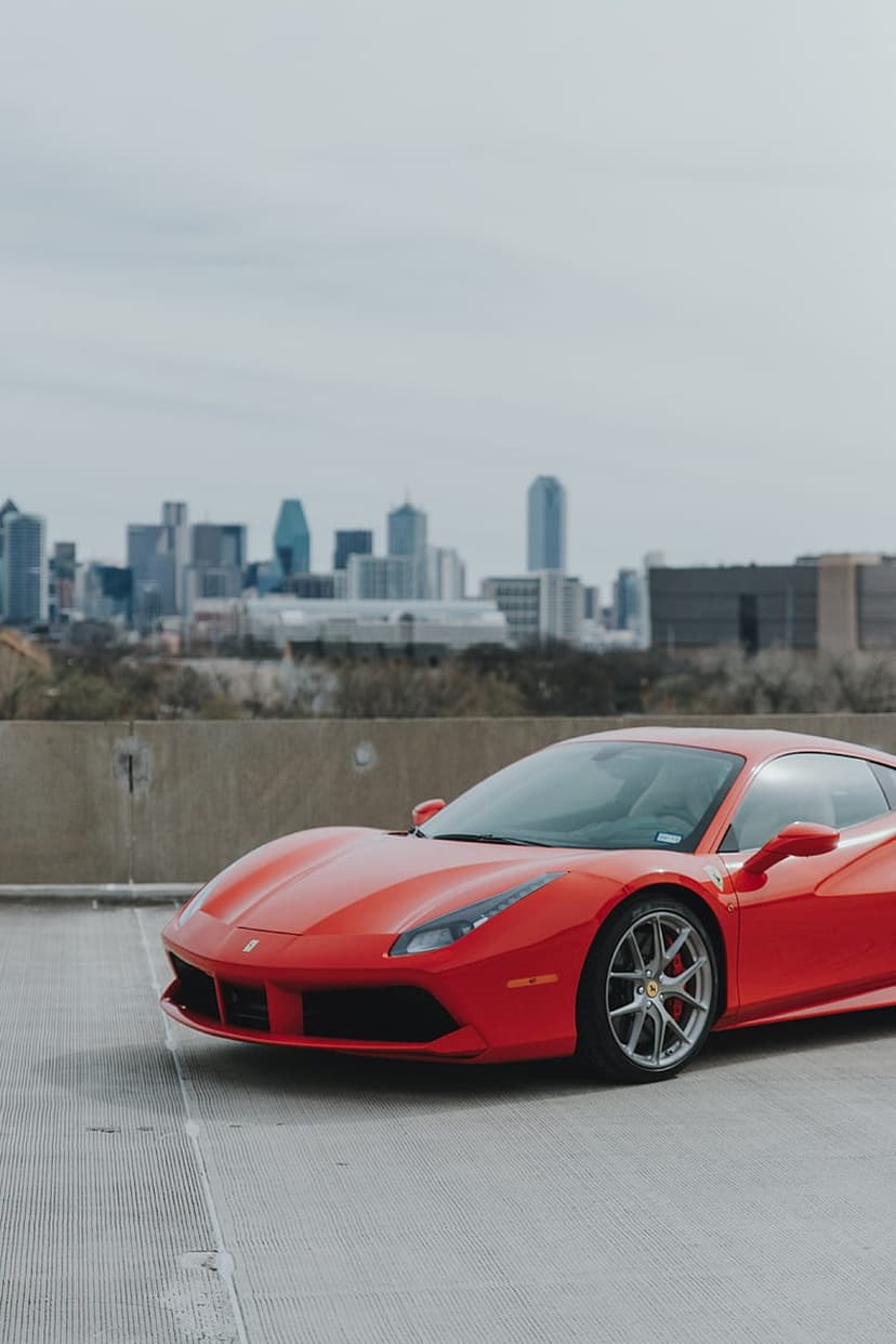 Red Ferrari sports car parked on a concrete lot with a blurred metropolitan skyline and skyscrapers behind it.