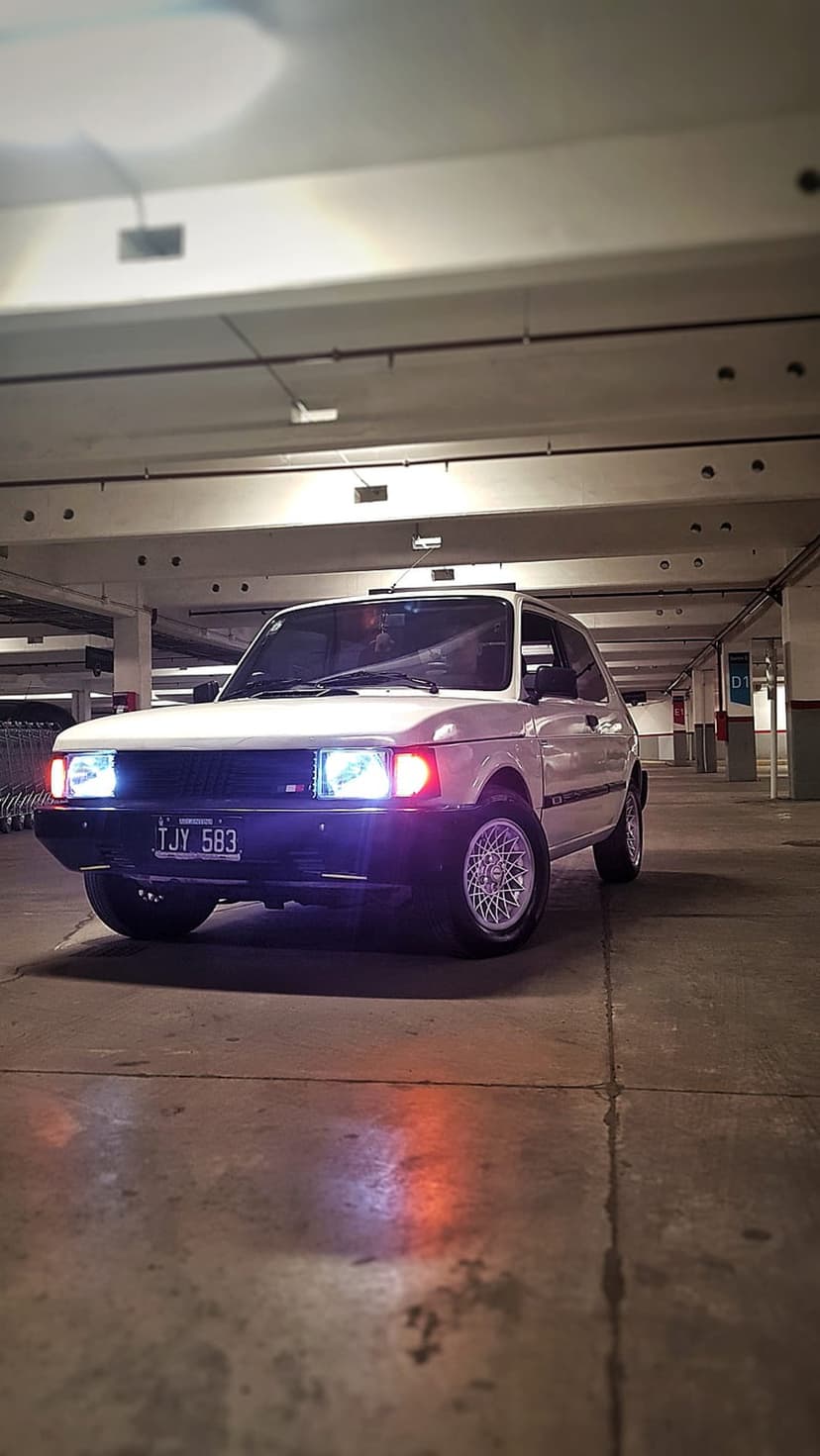 White Fiat 147 hatchback with glowing headlights in a dark parking garage with blue and purple floor reflections.