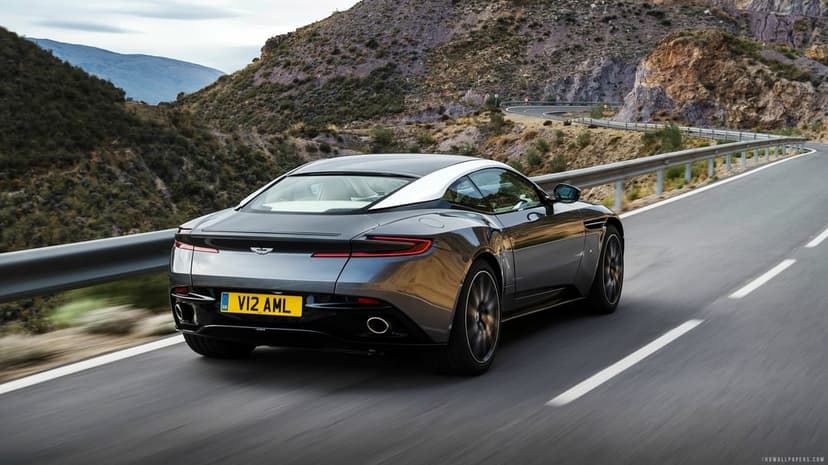 Grey Aston Martin DB11 driving on a winding mountain road with motion blur and rocky hill background.
