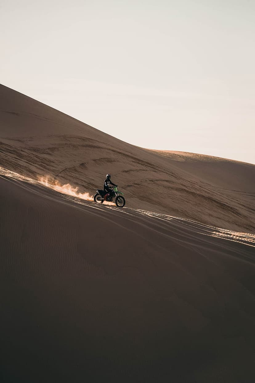 Motorcyclist riding down a sand dune at sunset with golden light and a dust trail in a vast desert landscape.