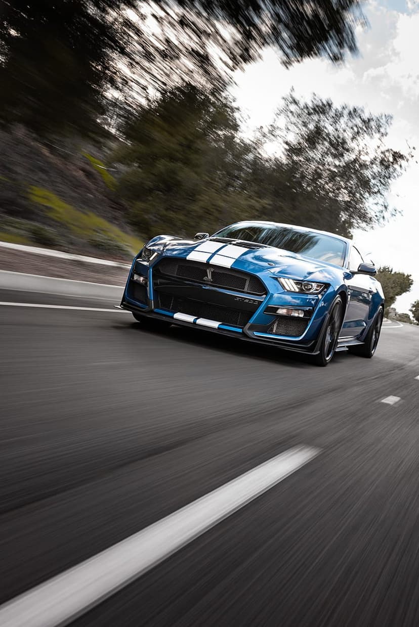 Blue Shelby GT500 Mustang with white stripes drives on a winding road bordered by thick green trees and foliage.