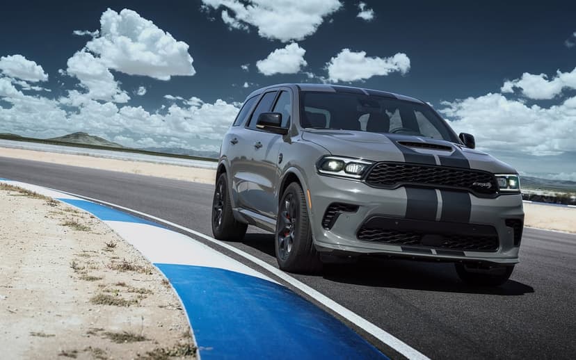 Grey Dodge Durango SRT Hellcat with black racing stripes driving on a curved racetrack under a bright blue sky.