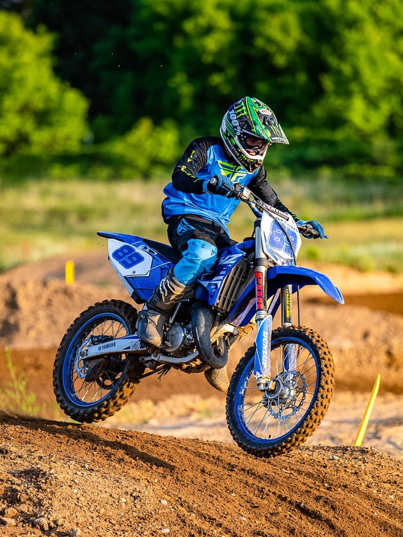 Motocross rider in blue gear jumping a Yamaha dirt bike over a dusty track with green trees in the background.
