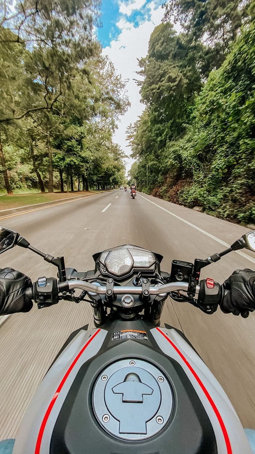 Motorcycle handlebars and a paved road stretching toward distant riders under a blue sky with fluffy clouds.