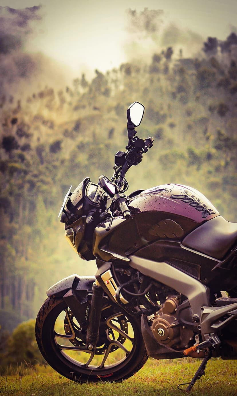 Dark motorcycle with rain droplets parked on green grass facing misty mountains and a dense evergreen forest.