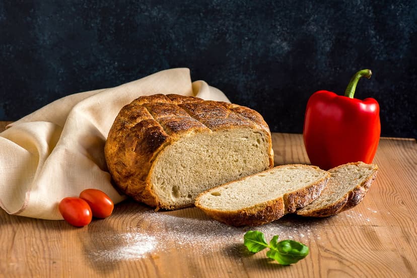 Sliced sourdough bread sits on a floured wood surface with a red bell pepper, cherry tomatoes, and a basil leaf.