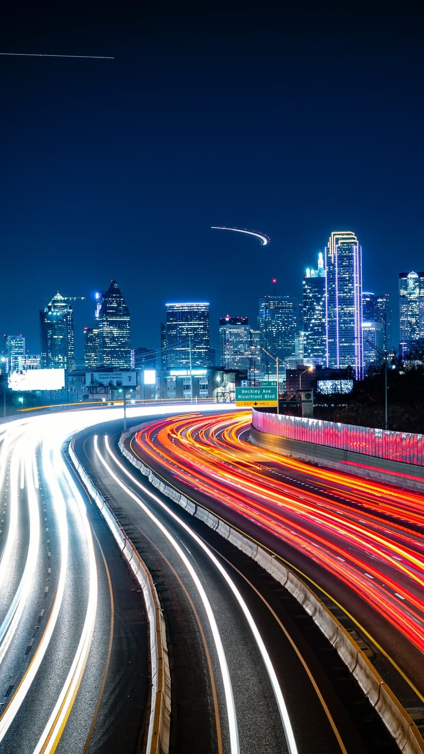 Red and white light trails streak along a city highway between illuminated skyscrapers under a dark night sky.