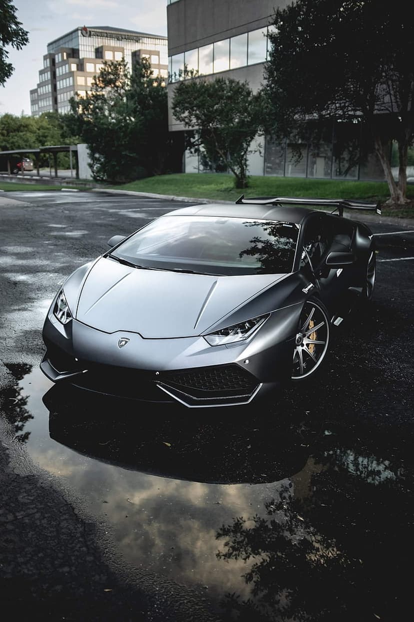 Silver Lamborghini Huracan with a rear wing parked on wet reflective asphalt near modern buildings and trees.
