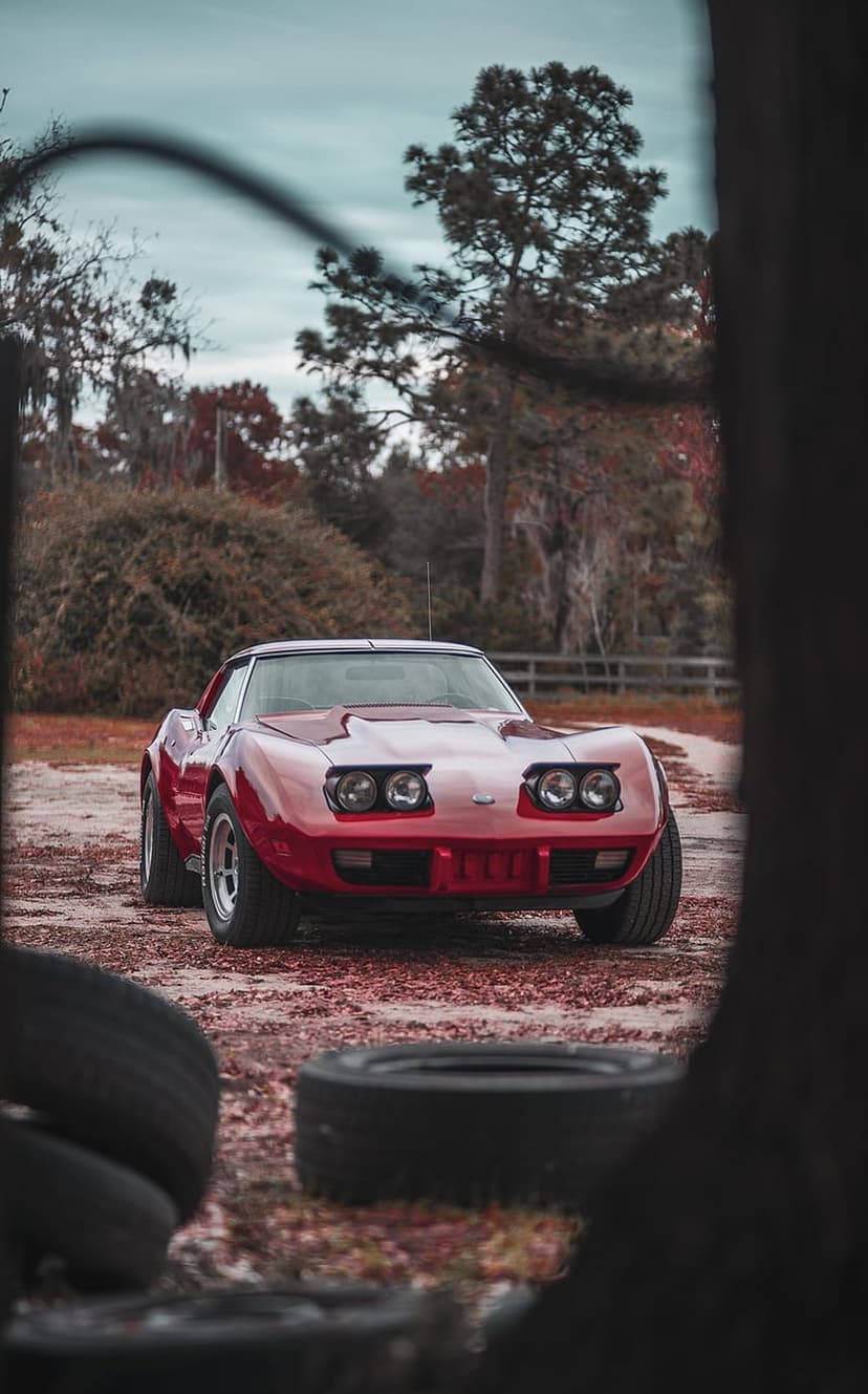 Red Corvette C3 parked on a dirt road with autumn leaves, pop-up headlights, and forest trees in the background.