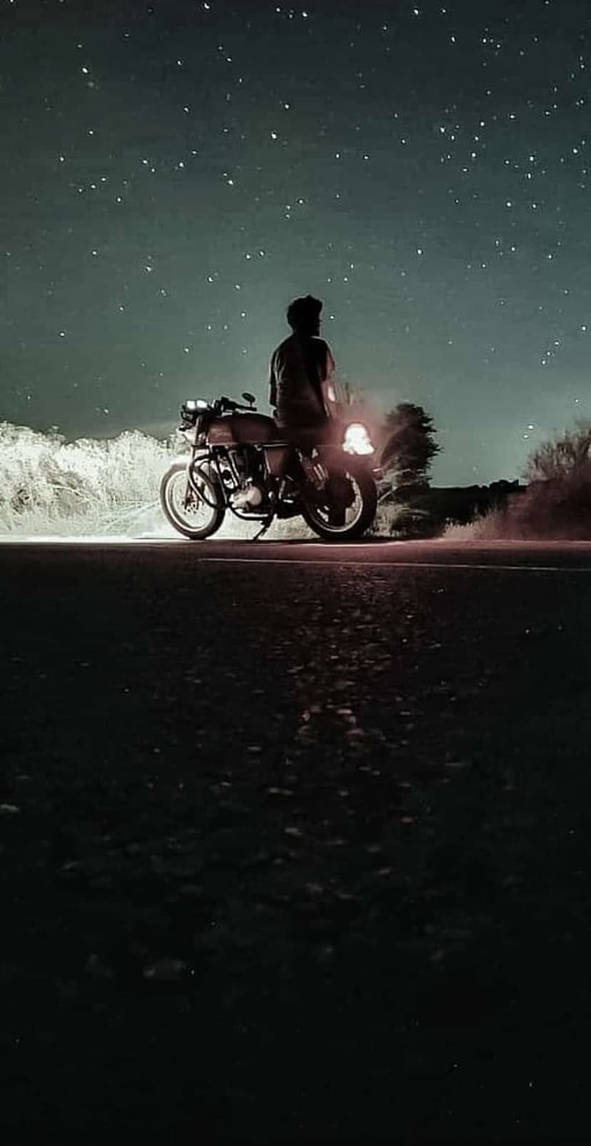 Motorcycle rider sitting on a bike beneath a vast starry sky with a bright headlight illuminating a dark road.