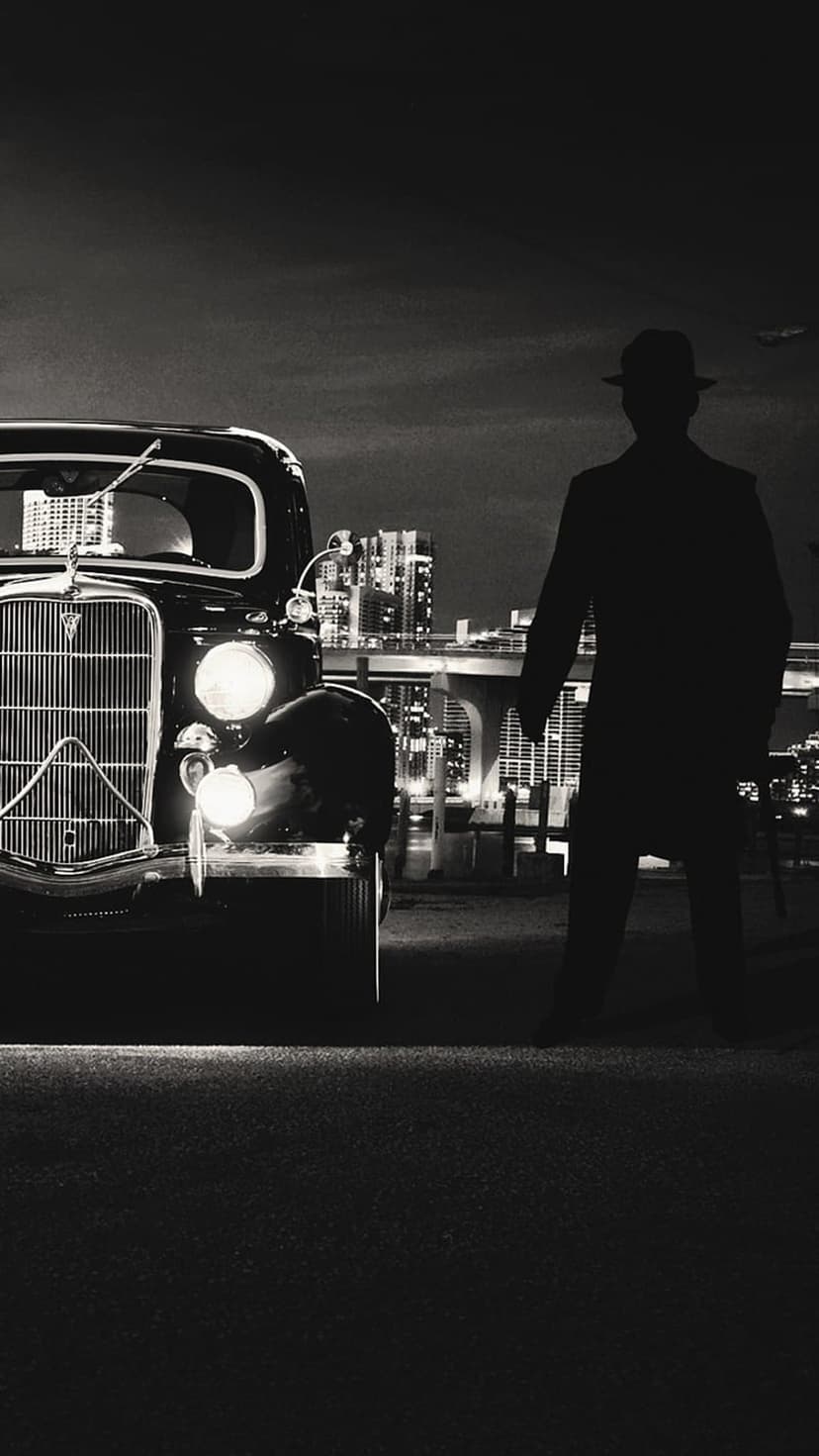 Man in a trench coat and fedora stands by a vintage car with glowing headlights against a dark city backdrop.