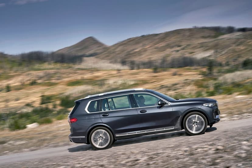 A dark gray BMW X7 SUV speeds along a gravel road against a backdrop of blurred rolling hills and dry grass.