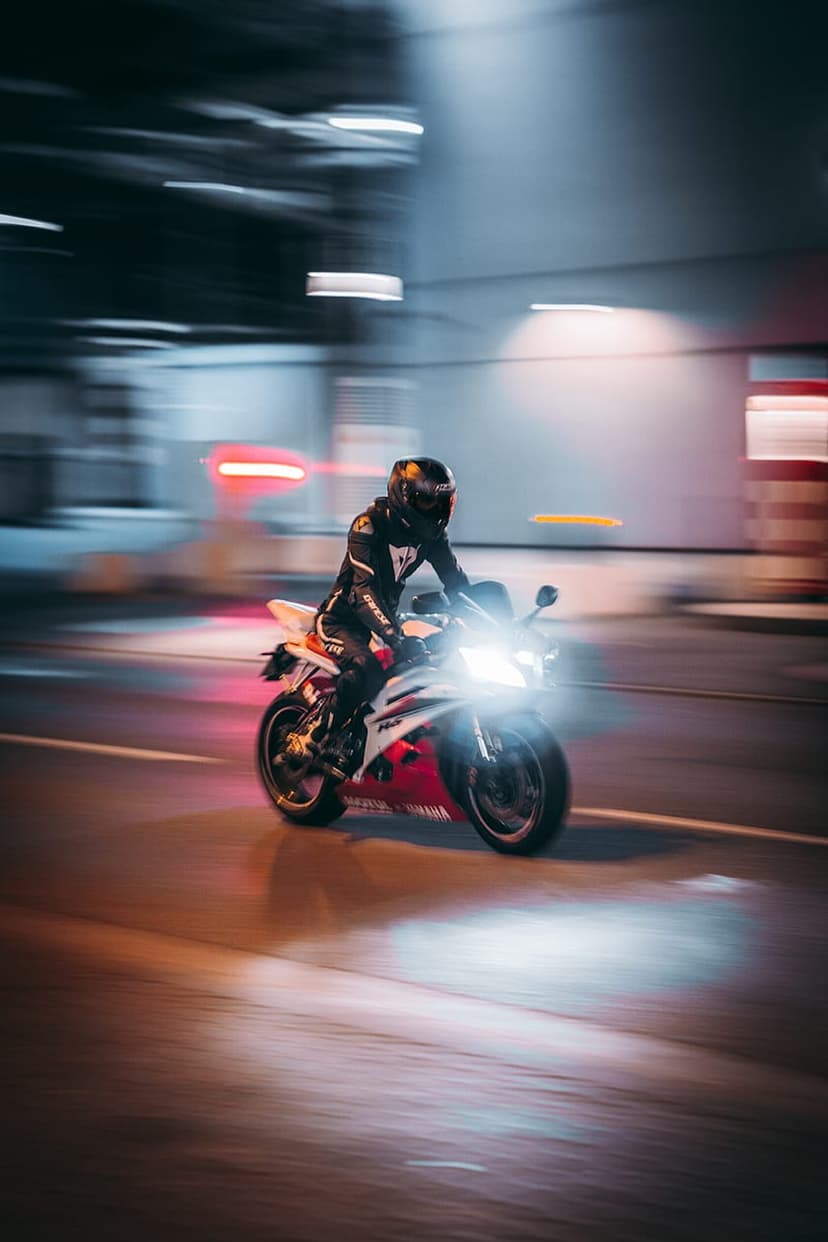 Motorcyclist in full gear leans on a sportbike as blurred city lights trail behind on a dark urban street.