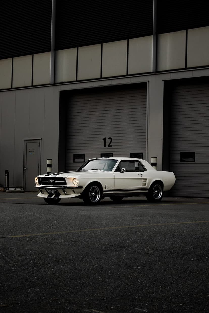 White 1960s Ford Mustang with black stripes parked on asphalt by an industrial building with garage doors.