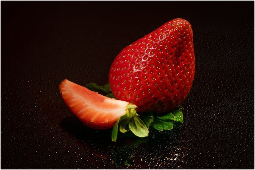 Red strawberry and a fresh slice on green leaves over a dark textured surface with clear water droplets.